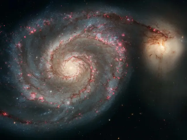 A spiral galaxy seen from overhead, with its two major arms spiraling out from its bright white core. At the end of the arm on the right is another bright white blob, this is a second galaxy. The arms are bluish and purple, peppered with countless stars.