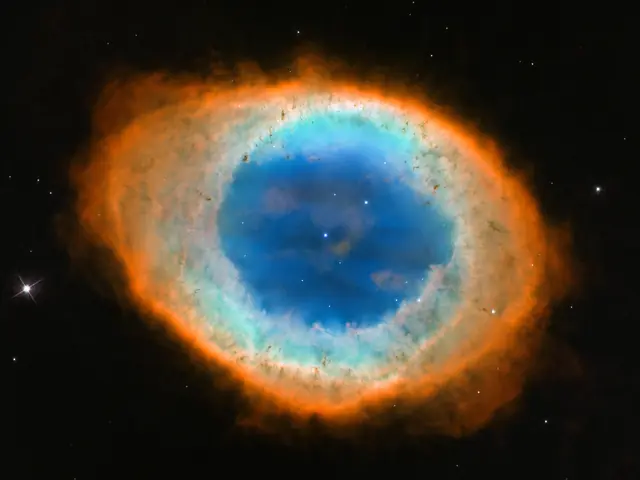 Black background holding an oval of light. The outer ring of the oval is orange-red. Moving inward the ring is more greenish-yellow. The center of the ring is bright blue with one small star at its center and another above and to the right of it.