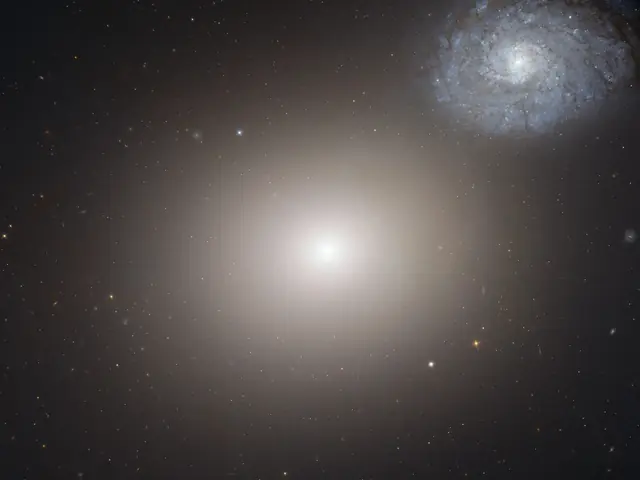 A hazy white-yellow light shines in the center of the image against black space, dotted with faint, distant stars. In the top right of the image is a spiral galaxy with many arms as seen from overhead. It is light blue.