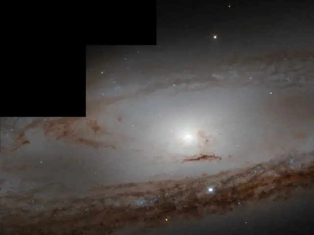 A spiral galaxy with a bright core fills the image, surrounded by faint spiral arms laced through with dark dust. A black rectangle is at the upper left, showing a portion where there is no Hubble data.