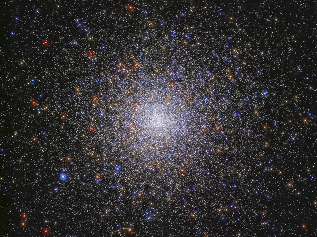 Image center holds a dense sphere of stars in colors of red, orange, yellow, white, and blue. The stars are densely packed at image center and taper out toward the image's edges. They are on a black background.