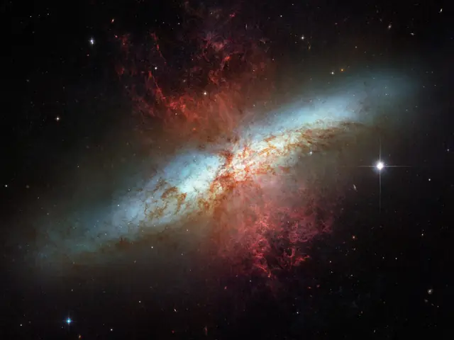 A white band of stars that cuts across a black background, from the lower left to the upper right, is the galaxy M82. Reddish brown gas and dust overlays the galaxy concentrated in the center of the image and fanning out above and below the white band of the galaxy. Black background is dotted with stars.