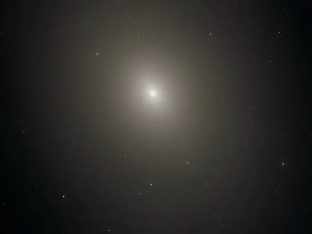 Diffuse oval haze against black background. Stars and distant galaxies shine through it. Just above image center the haze gets brighter and denser toward the bright-white core of the galaxy. Top, just left of center is a bright foreground star.