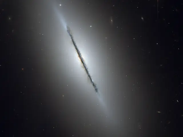 A galaxy is seen edge-on, with a dark lane of dust obscuring it surrounded by a hazy white glow, all against a black background.