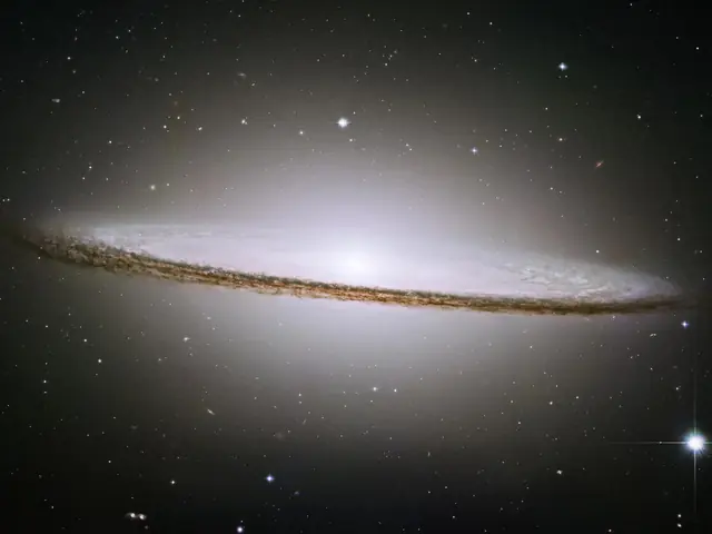 A bright hazy, oblate ball of stars fills the scene. Its core is bright white. Around the equator is a disk of bright stars and dark dust. The dust lane is prominent and bisects the sphere of stars.